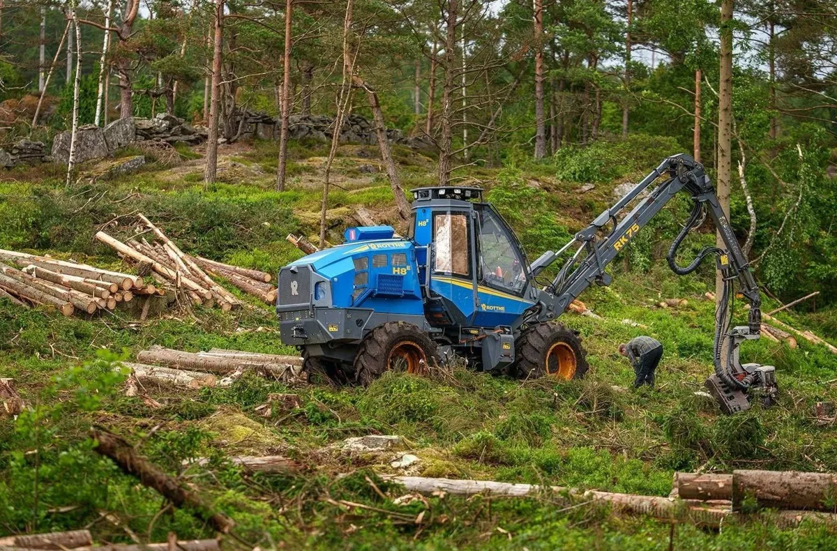 Blå Rottne H8-skördare gallrar i en skogsbacke med staplat virke